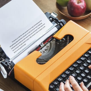 Fingers typing on a vintage orange typewriter near to a bowl of apples.